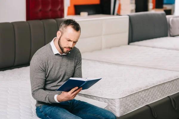 Hombre enfocado leyendo notas en cuaderno en tienda de muebles con colchones ortopédicos arreglados - foto de stock