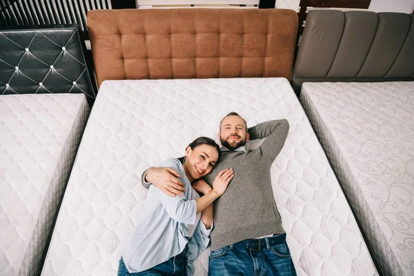 Overhead view of smiling couple lying on bed in furniture store with arranged mattresses — Stock Photo