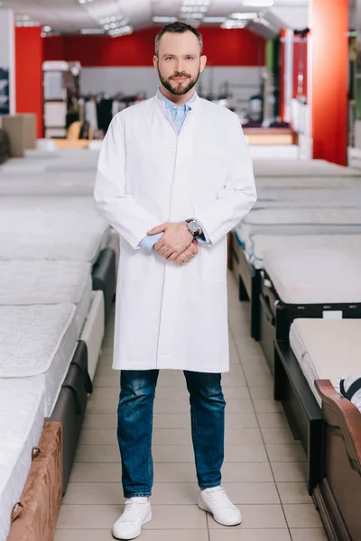 Shop assistant in white coat standing in furniture shop with arranged mattresses — Stock Photo