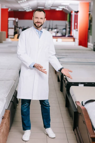 Smiling shop assistant pointing at orthopedic mattresses in white coat in furniture shop — Stock Photo