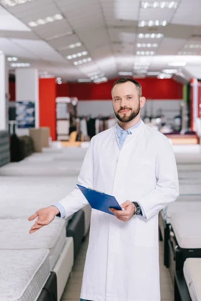 Shop assistant with notepad pointing at orthopedic mattresses in white coat in furniture shop — Stock Photo