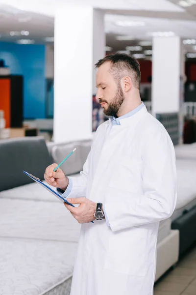 Side view of shop assistant in white coat making notes in notepad in furniture shop with arranged mattresses — Stock Photo