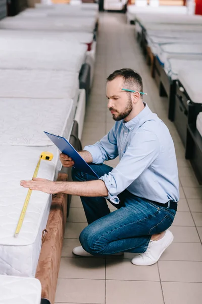 Shop assistant with notepad and measuring mattress with measure tape in furniture shop — Stock Photo