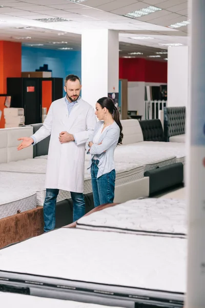 Male shop assistant in white coat showing goods to customer in furniture shop — Stock Photo