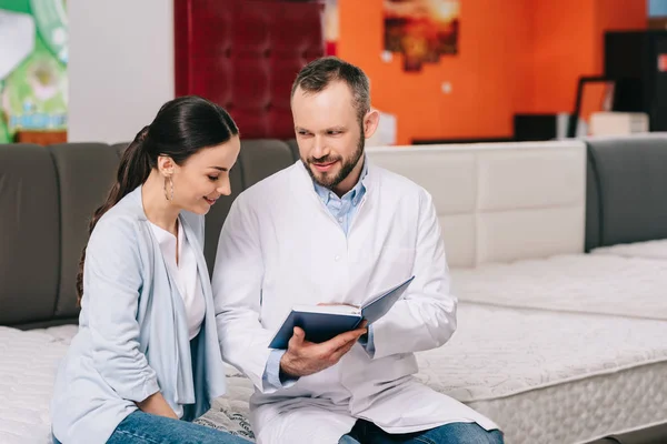 Male shop assistant in white coat with notebook helping woman in choosing mattress in furniture shop — Stock Photo