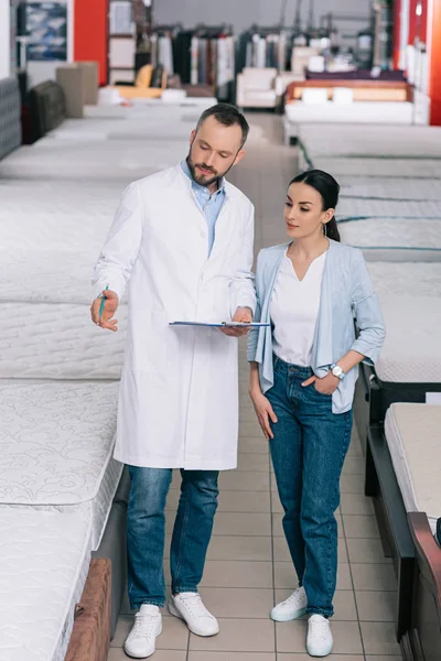 Male shop assistant in white coat with notebook helping woman in choosing mattress in furniture shop — Stock Photo