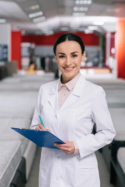 Focus selettivo di assistente negozio sorridente in cappotto bianco con blocco appunti in negozio di mobili con materassi disposti — Foto stock