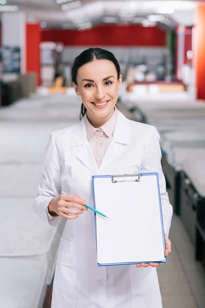 Assistente di negozio sorridente in cappotto bianco che indica blocco note vuoto in mano nel negozio di mobili con materassi disposti — Foto stock