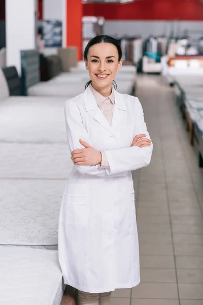 Portrait of smiling shop assistant in white coat with arms crossed in furniture shop with arranged mattresses — Stock Photo
