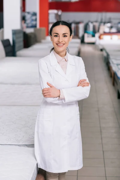 Retrato de asistente de tienda sonriente en bata blanca con brazos cruzados en tienda de muebles con colchones arreglados - foto de stock