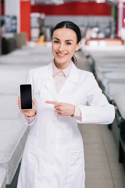 Asistente de tienda sonriente en bata blanca que muestra el teléfono inteligente con pantalla en blanco en la mano en la tienda de muebles con colchones - foto de stock