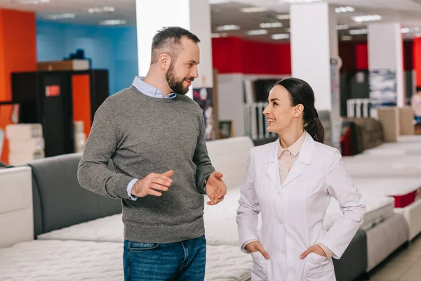 Foyer sélectif du client masculin et de l'assistant de magasin en manteau blanc dans le magasin de meubles avec matelas disposés derrière — Photo de stock