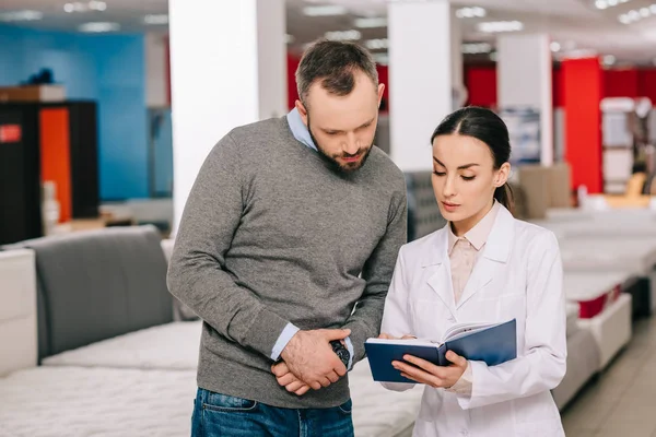 Female shop assistant in white coat with notebook helping customer in choosing mattress in furniture store — Stock Photo