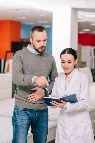 Female shop assistant in white coat with notebook helping customer in choosing mattress in furniture store — Stock Photo