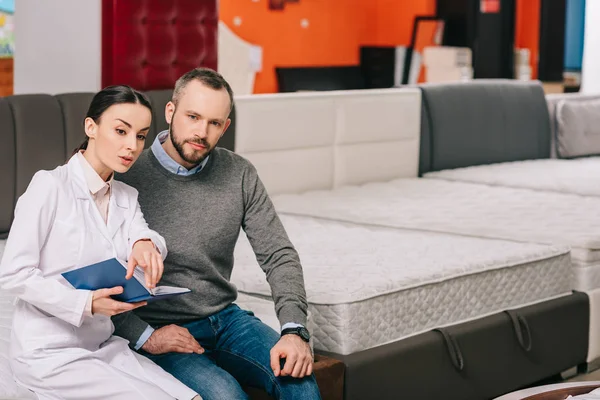 Commessa donna in cappotto bianco con taccuino che aiuta il cliente nella scelta del materasso nel negozio di mobili — Foto stock