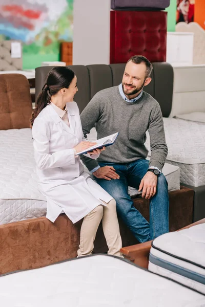 Female shop assistant in white coat with notebook helping customer in choosing mattress in furniture store — Stock Photo