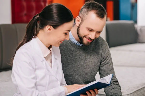 Assistente di negozio femminile sorridente in cappotto bianco con notebook aiutare il cliente nella scelta del materasso nel negozio di mobili — Foto stock