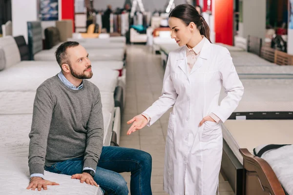 Messa a fuoco selettiva di cliente maschio e commessa in camice bianco in negozio di mobili con materassi disposti dietro — Foto stock