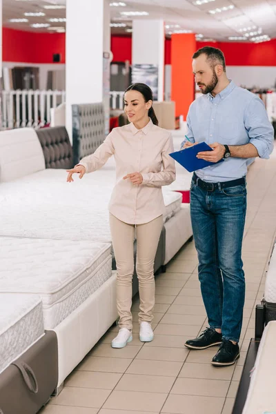 Couple with notepad choosing mattress together in furniture store with arranged mattresses — Stock Photo