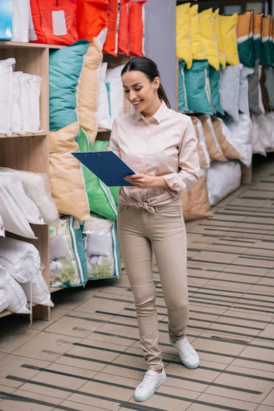 Assistente de loja sorridente com bloco de notas na loja de móveis com travesseiros arranjados — Fotografia de Stock