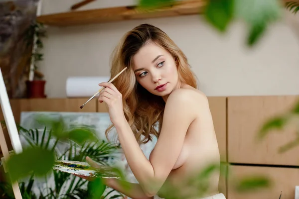 Selective focus of naked young female painter holding brush with palette and looking away in art studio — Stock Photo