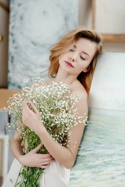 Sensual naked girl with closed eyes holding white flowers and leaning at easel in art studio — Stock Photo