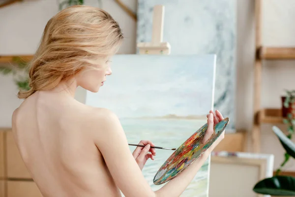 Back view of naked young artist holding brush and palette in art studio — Stock Photo