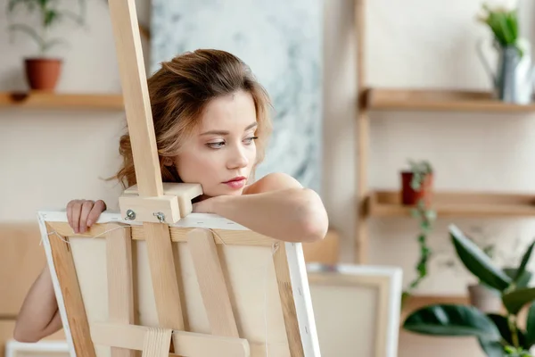 Beautiful sensual young woman leaning at easel and looking away in art studio — Stock Photo