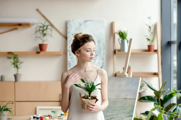 Hermosa tierna artista femenina sosteniendo planta en maceta y mirando hacia otro lado en el estudio de arte - foto de stock