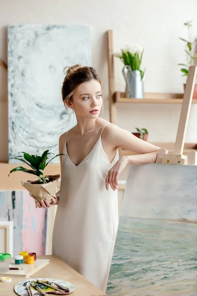 Hermosa artista joven con maceta verde planta apoyada en la imagen en caballete y mirando hacia otro lado en el estudio de arte - foto de stock