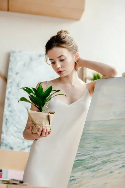 Schöne junge Künstlerin lehnt an Bild auf Staffelei und betrachtet grüne Topfpflanze — Stockfoto