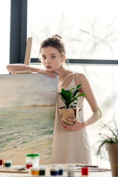 Hermosa joven pintora sosteniendo maceta planta y apoyándose en caballete en estudio de arte - foto de stock