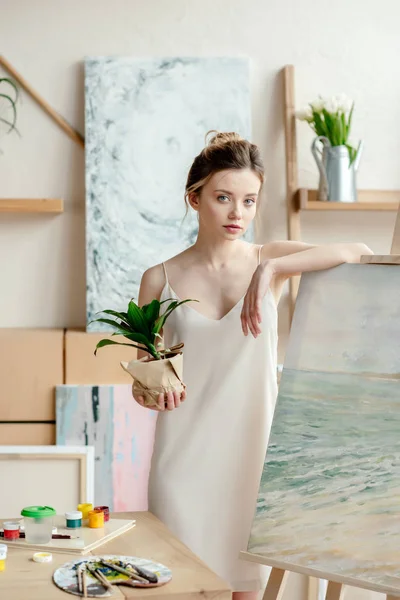 Bela menina macia segurando vaso planta e inclinando-se no cavalete no estúdio de arte — Fotografia de Stock