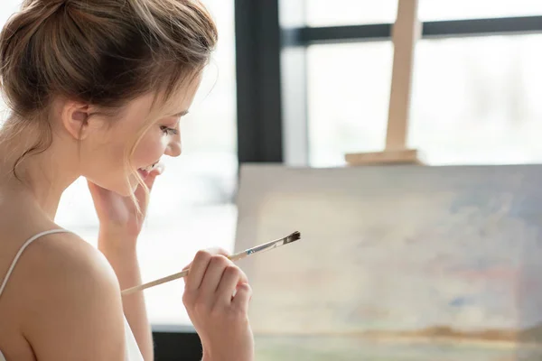 Beautiful smiling young woman holding brush and looking down in art studio — Stock Photo