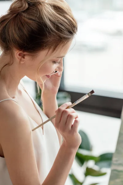 Beautiful smiling young painter holding brush and talking by smartphone in art studio — Stock Photo