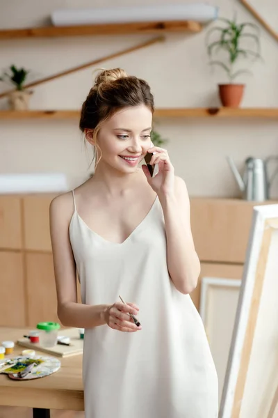 Beautiful smiling young painter holding brush while talking by smartphone and looking at easel in art studio — Stock Photo