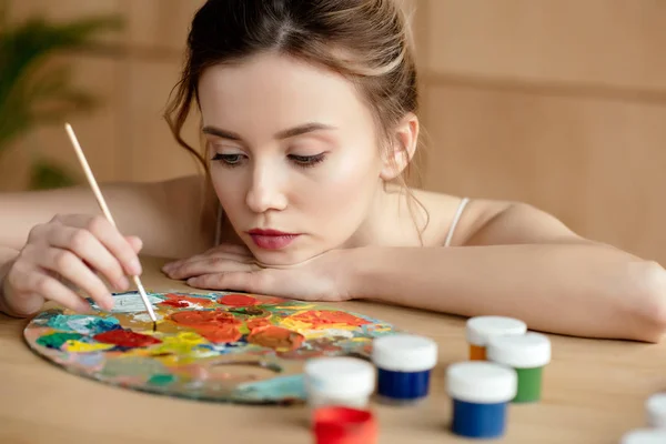 Beautiful young artist holding brush and looking at palette in art studio — Stock Photo