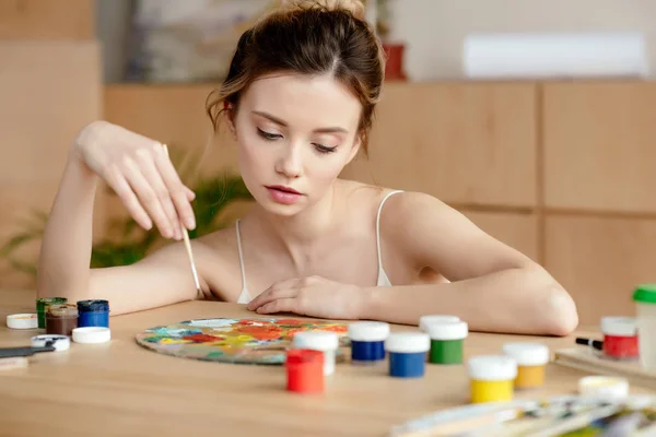 Hermosa joven pintora sosteniendo cepillo y mirando a la paleta en el estudio de arte - foto de stock