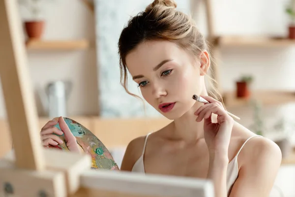 Attractive girl with brush and palette painting picture in art studio — Stock Photo