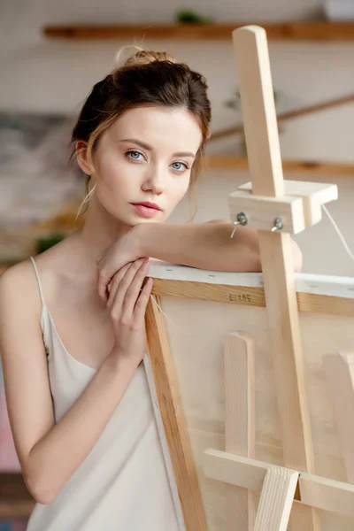 Schöne junge Frau lehnt an Bild auf Staffelei und blickt in Kamera im Kunstatelier — Stockfoto