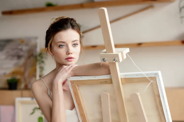 Beautiful pensive young artist leaning at easel and looking away — Stock Photo