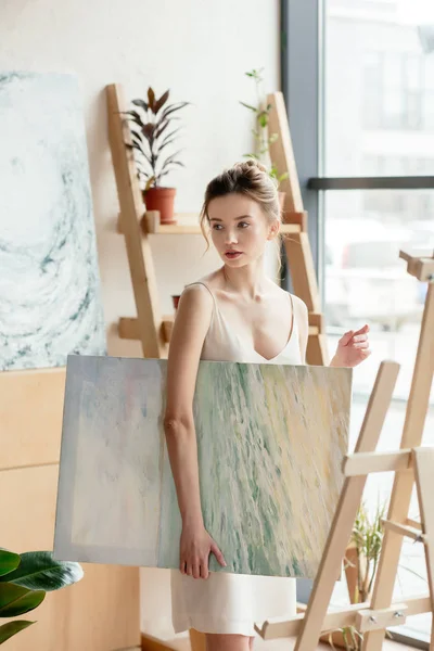Schöne junge Frau hält Bild in der Hand und schaut im Kunstatelier weg — Stockfoto