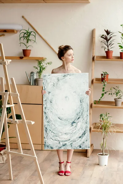 Full length view of beautiful young naked artist in red sandals holding picture and looking away in studio — Stock Photo