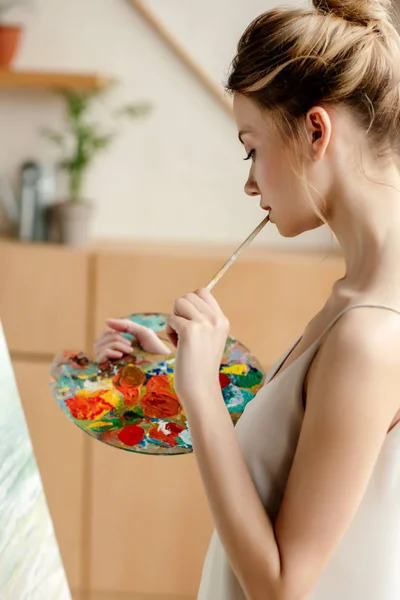 Vista laterale di attraente giovane artista con tavolozza e pittura a pennello su cavalletto in studio — Foto stock