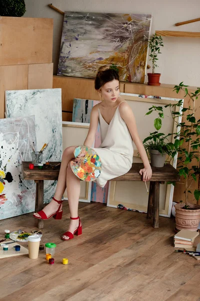 Hermosa joven artista elegante sosteniendo paleta y mirando hacia otro lado en el estudio - foto de stock