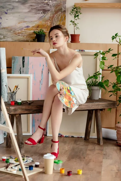 Schöne junge Künstlerin hält Palette und sitzt im Atelier — Stockfoto