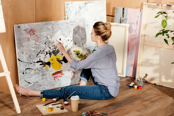 Beautiful young female painter sitting on floor and drawing picture in art studio — Stock Photo