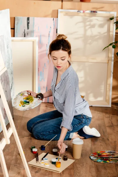 Beautiful girl holding palette and painting in art studio — Stock Photo