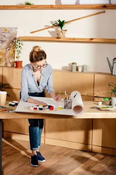 Stylische junge Brillenkünstlerin malt am Tisch im Atelier — Stockfoto