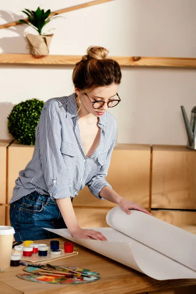 Stylish female artist in eyeglasses rolling canvas on table with painting supplies — Stock Photo